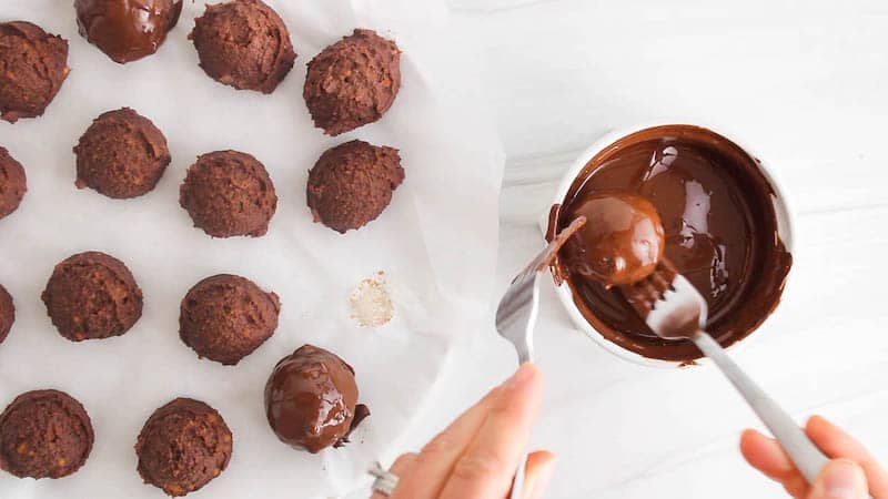 There are 2 forks dipping a chocolate chickpea truffle in melted chocolate.