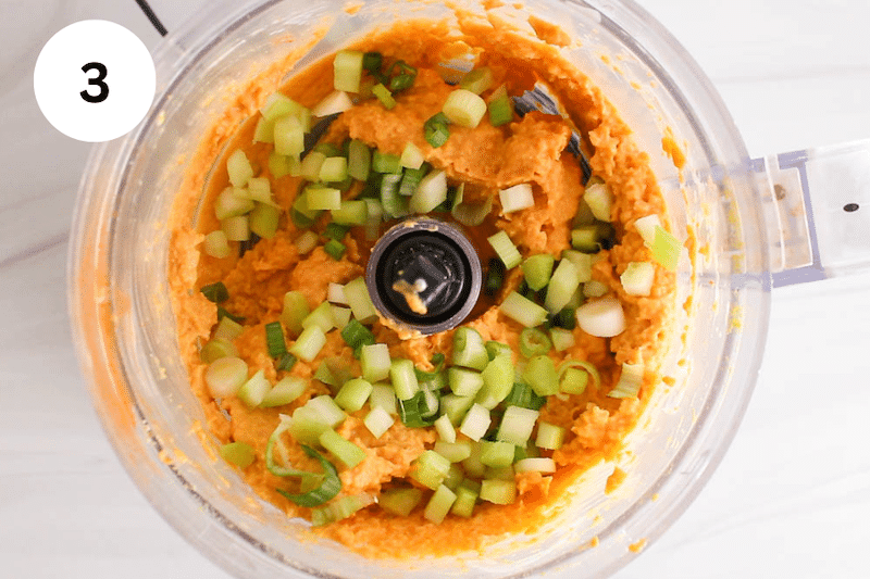 Green onion and celery pieces over a creamy orange mix in a food processor.