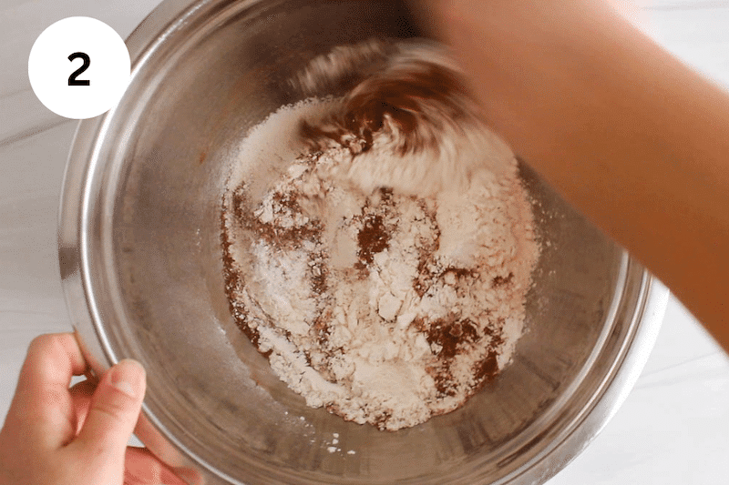 A large spoon stirring some flour, cinnamon, baking powder and salt in a large metallic bowl.