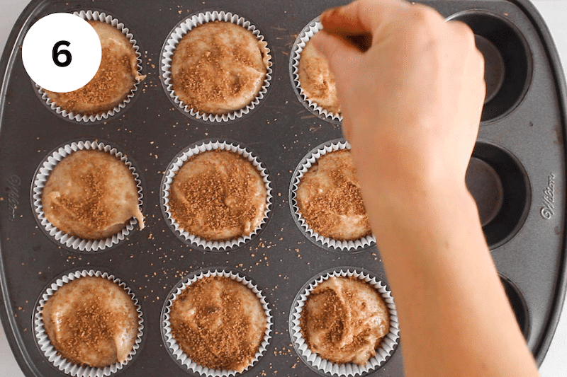 A hand is sprinkle cinnamon sugar over muffins in a muffin pan.