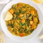 White bowl of sweet potato and spinach red lentil dhal garnished with cilantro and lemon wedges.