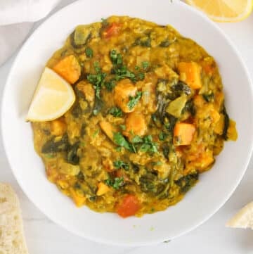 White bowl of sweet potato and spinach red lentil dhal garnished with cilantro and lemon wedges.