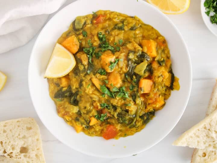 White bowl of sweet potato and spinach red lentil dhal garnished with cilantro and lemon wedges.