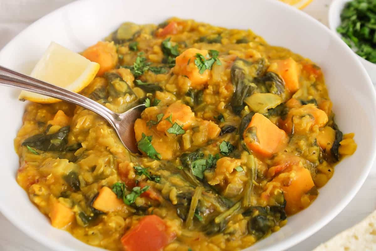 A spoon digging in a bowl of red lentil sweet potato dhal made with spinach.