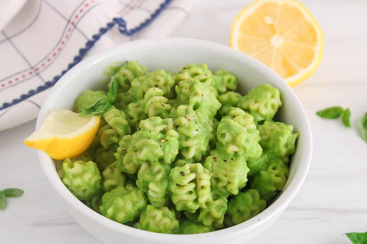 A white bowl filled with short pasta tossed in a green pea pasta sauce and garnished with black pepper.