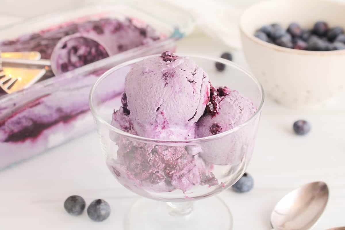 3 scoops of vegan blueberry ice cream in a see-through glass bowl with fresh blueberries in the background.