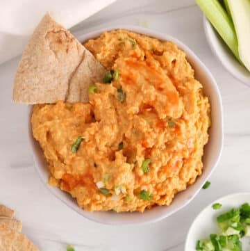 Vegan buffalo chickpea dip in a bowl with pita bread and raw veggie sticks.