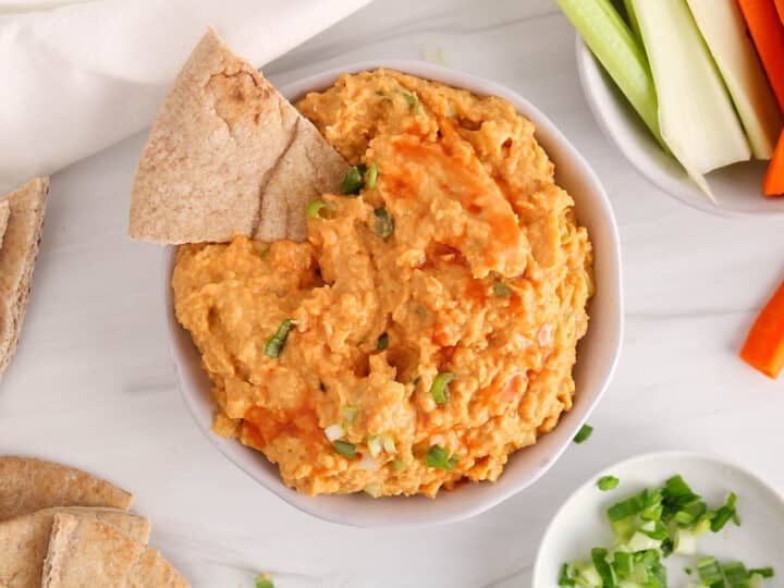 Vegan buffalo chickpea dip in a bowl with pita bread and raw veggie sticks.