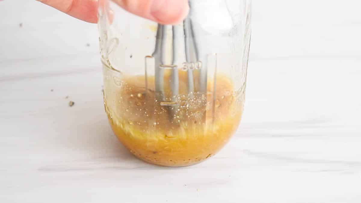 Small whisk stirring in a small jar of salad dressing.