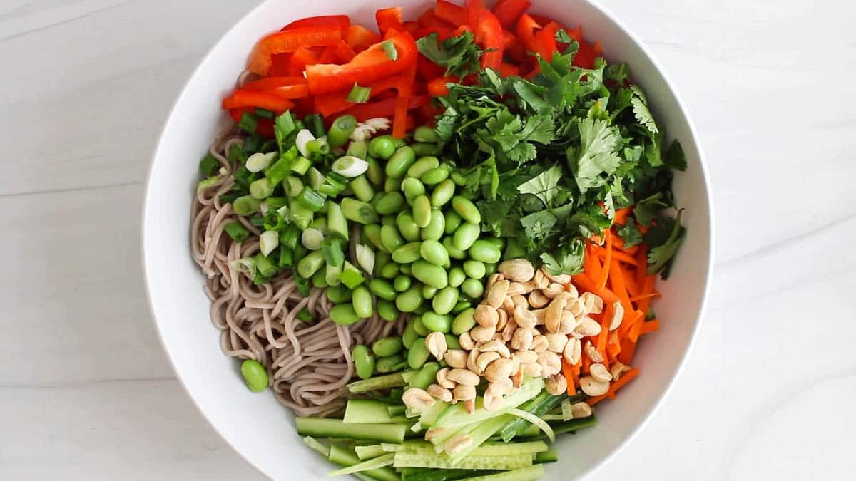 In a large bowl, there are noodles, fresh veggies, edamame and peanuts.