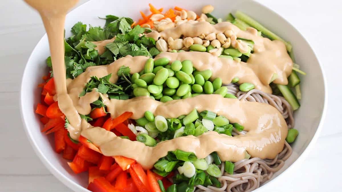Pouring a peanut butter sauce over a bowl of noodles and fresh veggies.