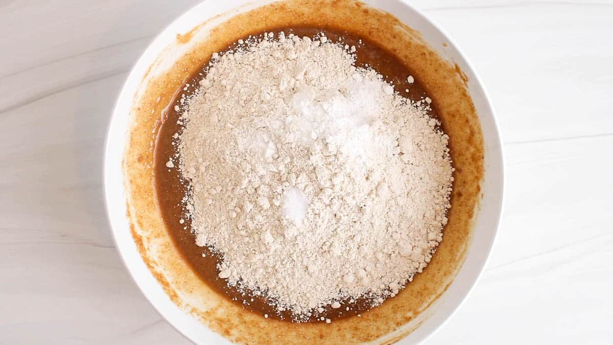 Flour over a wet brown cookie mixture in a white bowl.