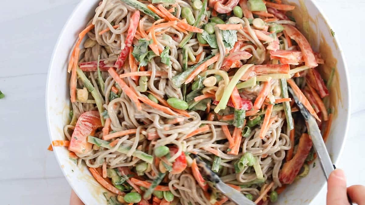 Tongs stirring noodles and veggies with a peanut sauce.