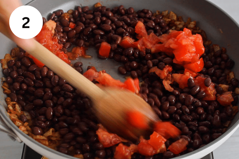 A wooden spoon stirring in a pan with tomatoes and black beans.