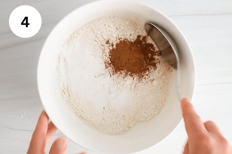 Flour, cinnamon and leavening agents in a white bowl with a large spoon.