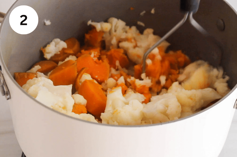 A potato masher mashing some sweet potatoes and cauliflower.