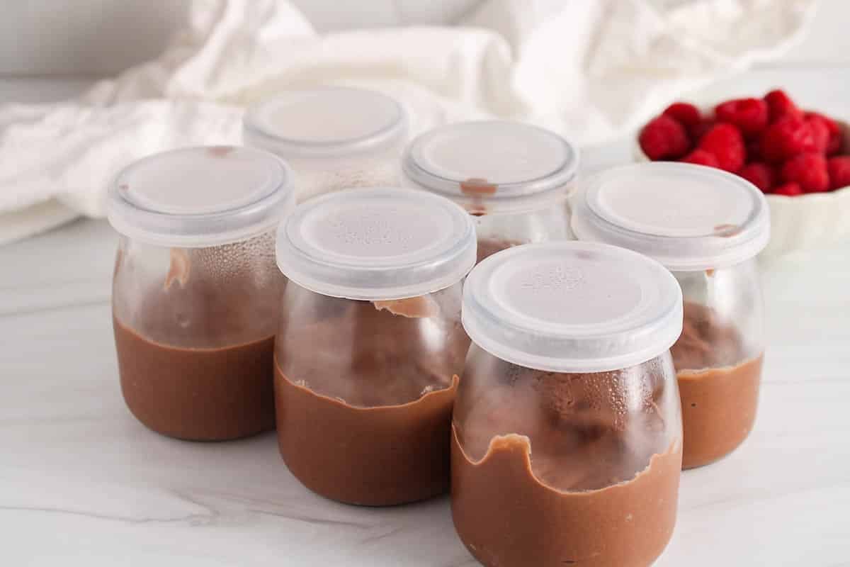 6 small jars of cashew chocolate mousse with a plastic cover on on a white table.