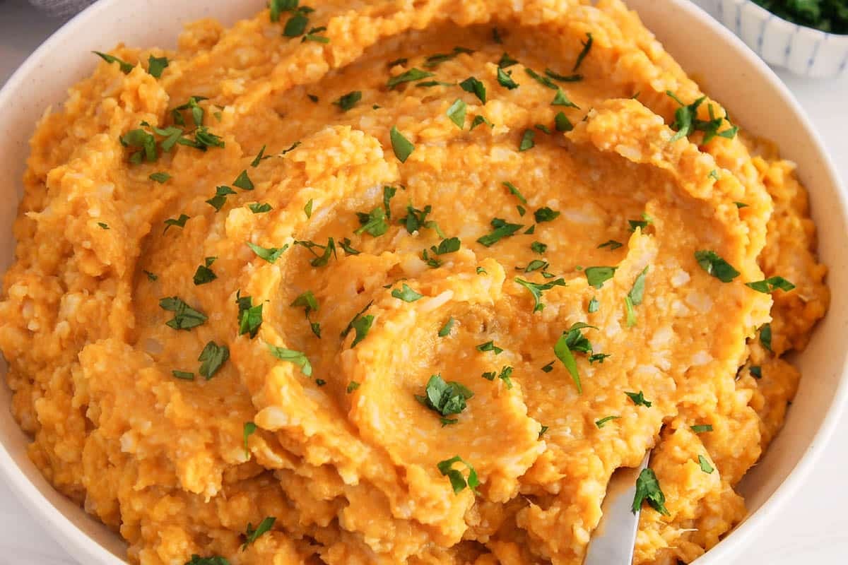 A spoon placed in a bowl of mashed sweet potato and cauliflower garnished with herbs.
