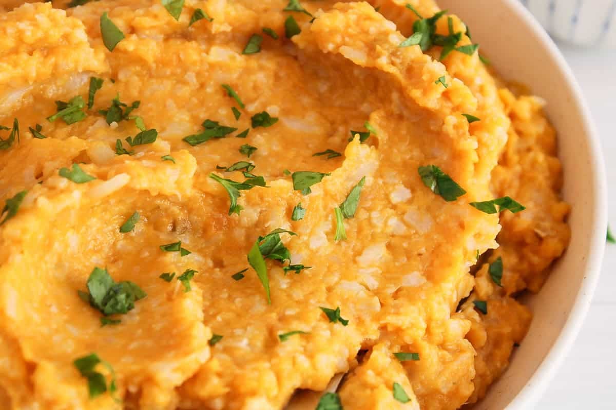 Coarse mash of sweet potato and cauliflower with parsley in a bowl with a spoon.