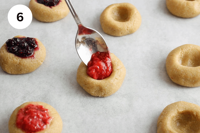A spoon adding jam in a thumbprint cookie.