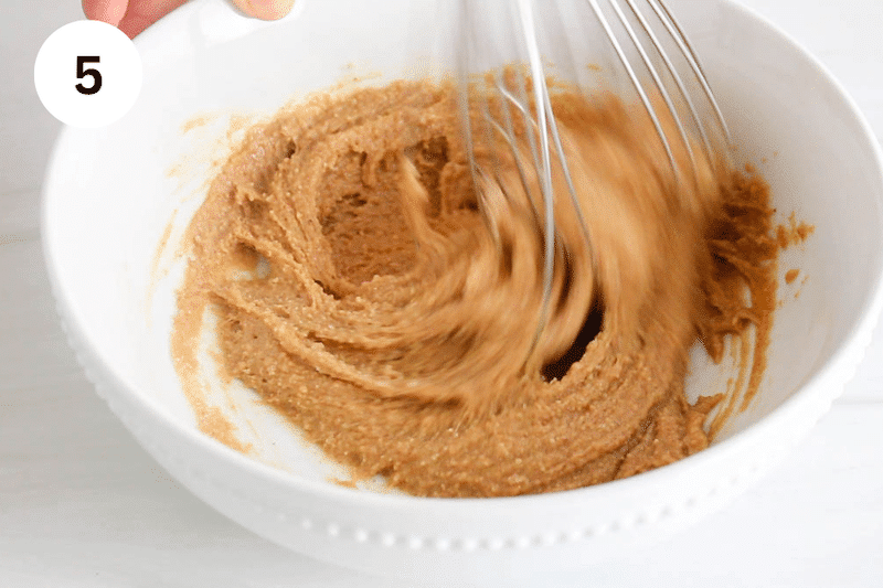 A large whisk stirring in a bowl with peanut butter and sugar.