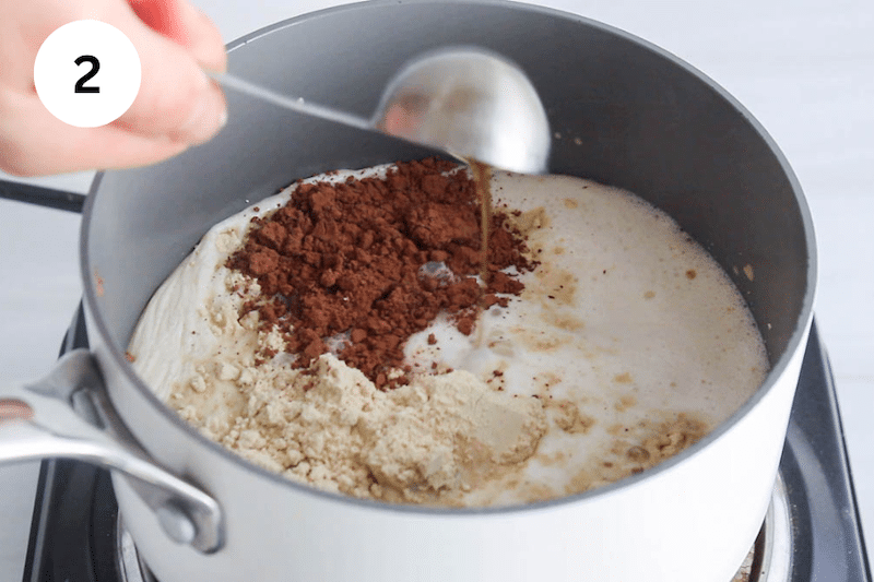 A small saucepan with milk, cacao powder and protein powder while a tablespoon is pouring some maple syrup.