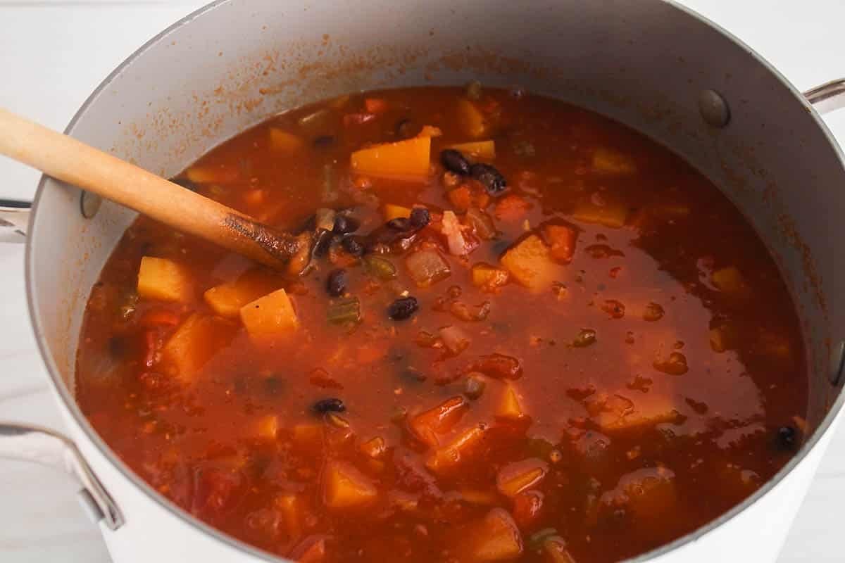 A large pot with a tomato, black bean and butternut squash soup with a wooden spoon stirring.