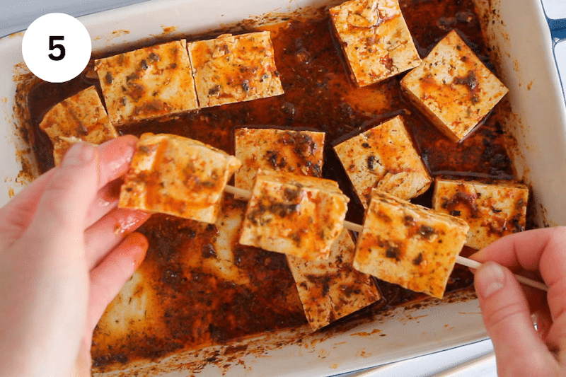 Hands transferring marinated tofu over a skewer.