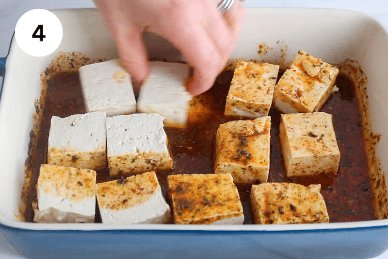 A hand stirring large cubes of tofu in a container with a marinade.