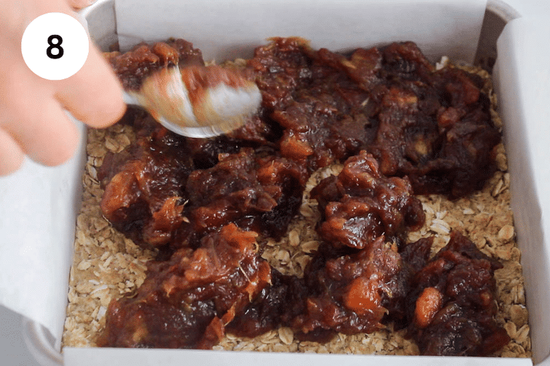 A spoon transferring a date mixture over a oat mix in a square pan.