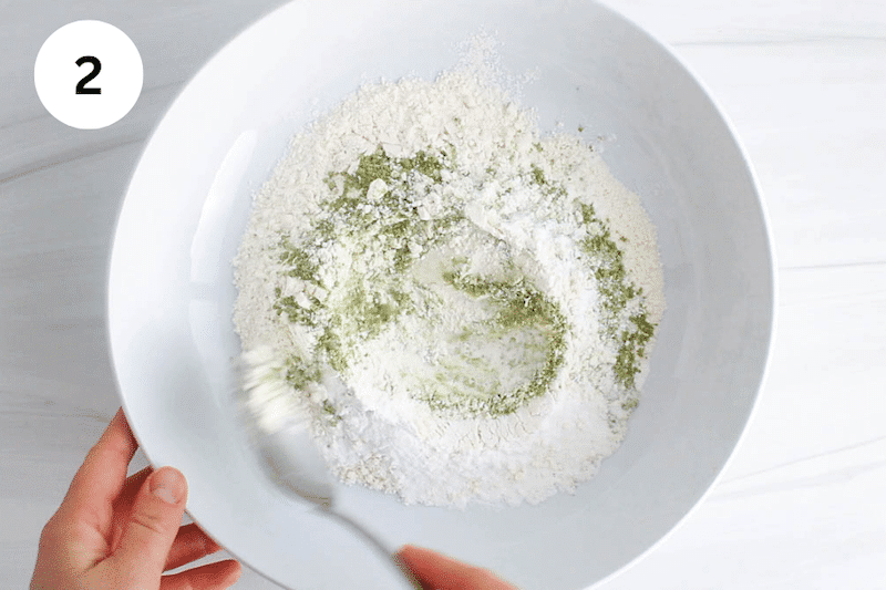 A white bowl with flour and matcha powder while a spoon is stirring.