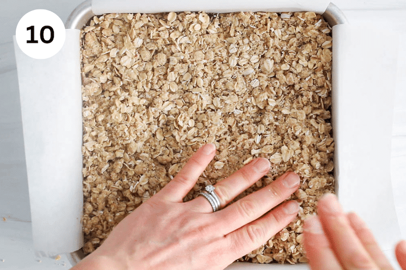 2 hands pressing over an oat mixture over some bars in a square pan.