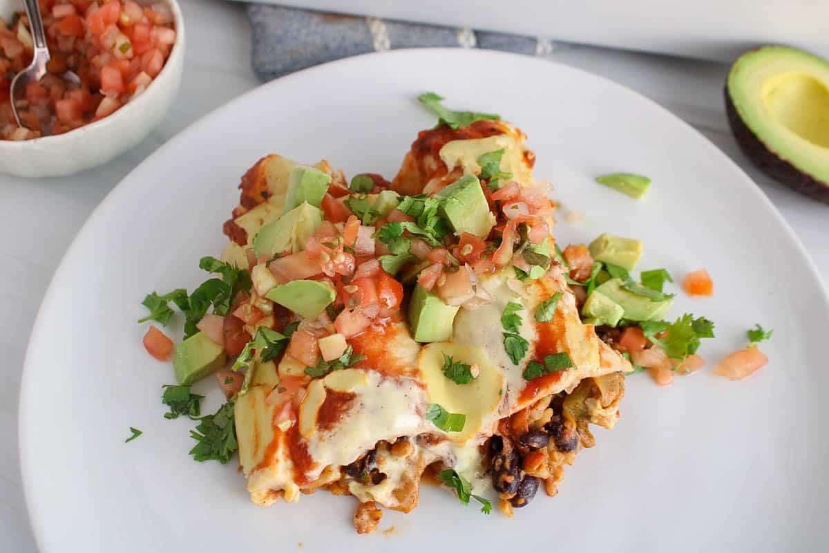 Two half of vegan bean and rice enchiladas on a serving plate garnished with avocado and pice de gallo.