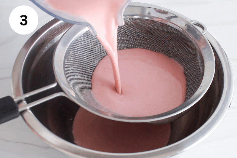 A pink strawberry oat milk pouring into a fine mesh colander over a large bowl.