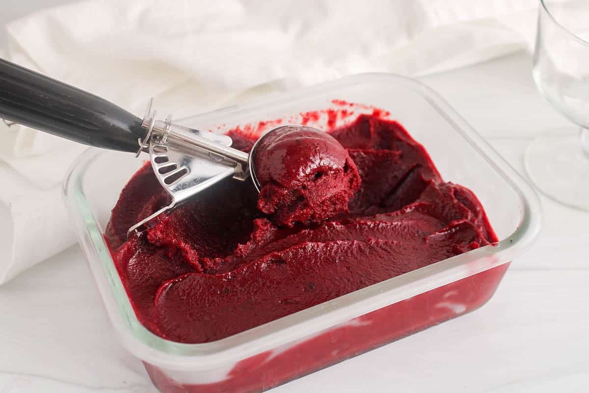 An ice cream scoop taking some cherry sorbet from a glass bowl.