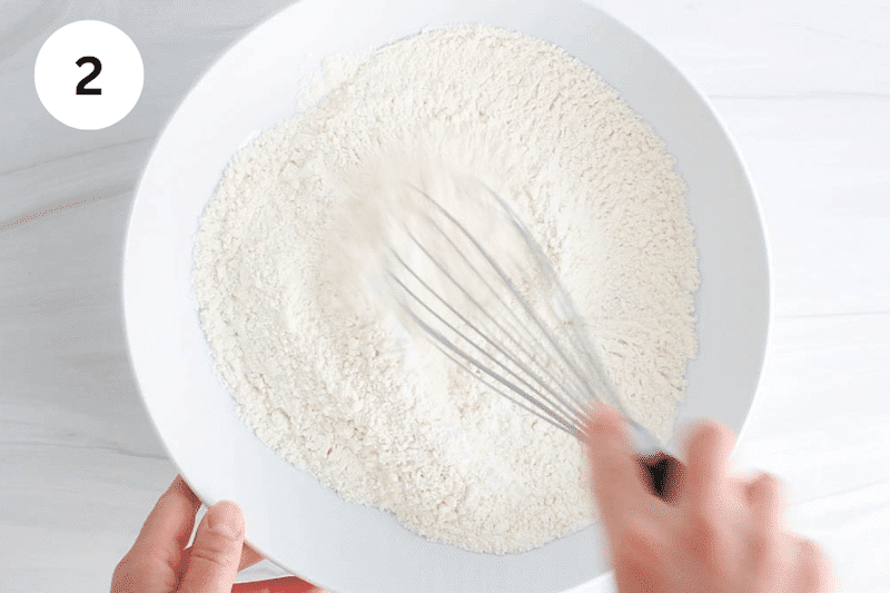 A large whisk mixing in a bowl with flour.