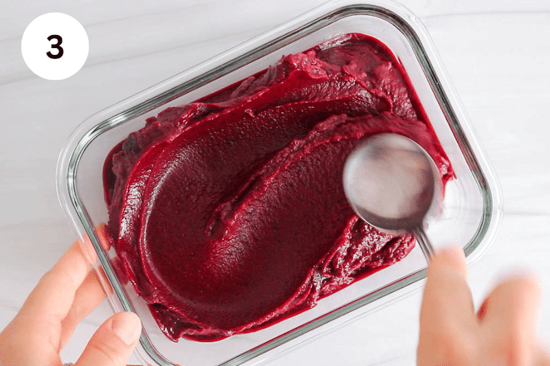 A spoon evening out the surface of a cherry sorbet in a glass container.