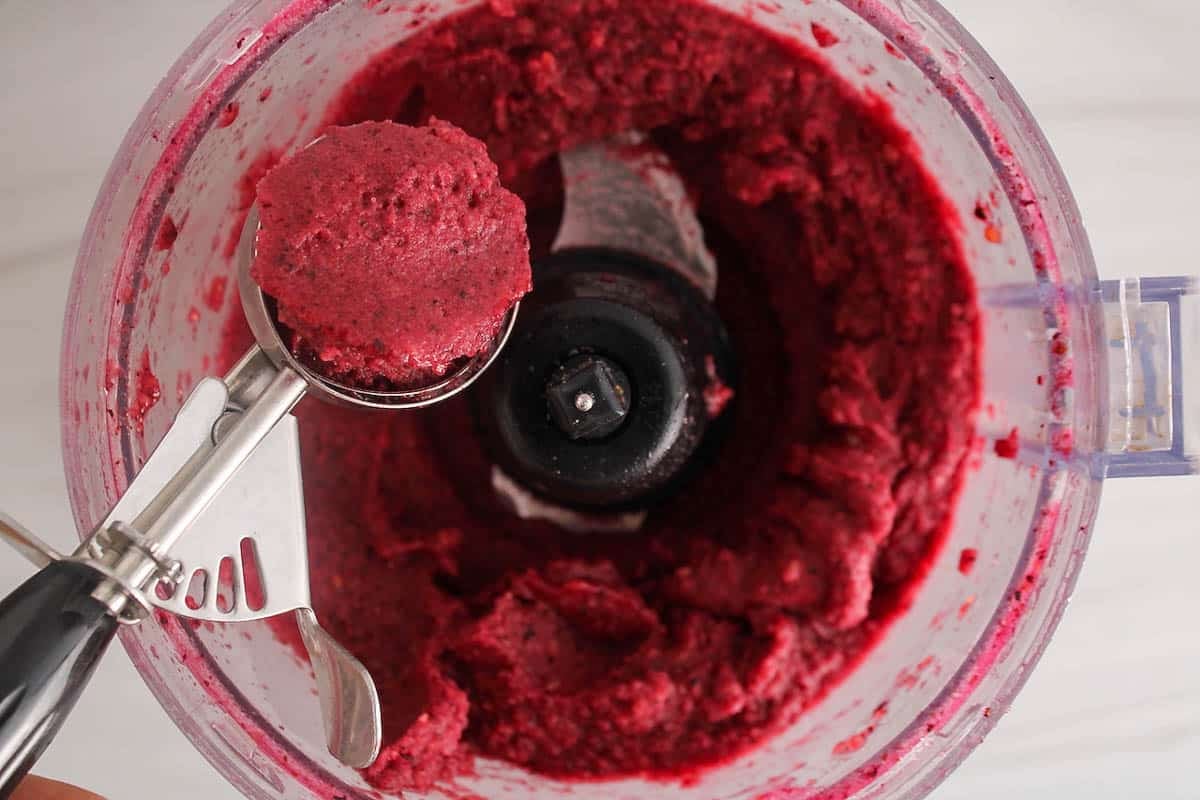 A food processor filled with homemade blueberry peach sorbet and an ice cream scoop taking some to serve.