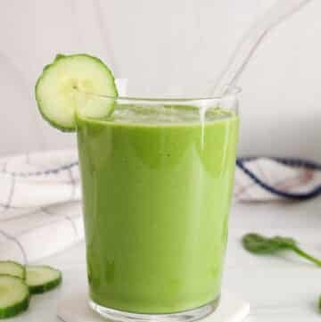 Tall glass of cucumber spinach smoothie with a straw and a slice of cucumber against the rim.