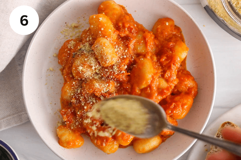 A spoon sprinkling some vegan parmesan cheese over red pesto gnocchi.