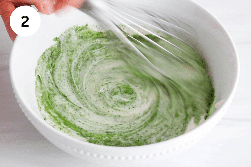A large whisk stirring a bowl of yogurt with matcha powder.