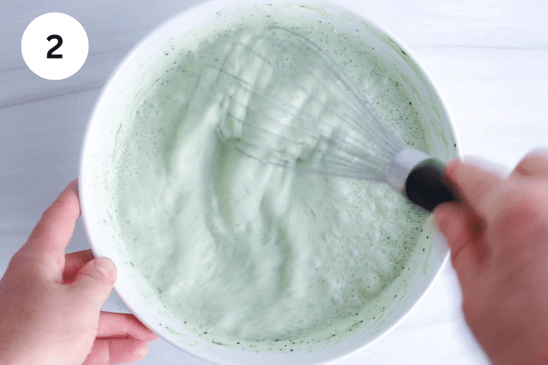 A large whisk stirring in a bowl with a green tea mix.