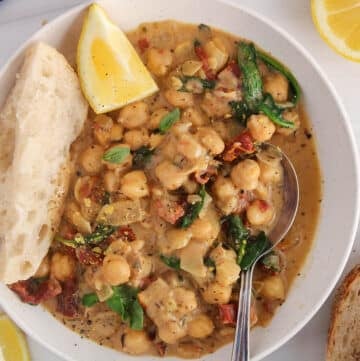 A spoon in a bowl of creamy vegan Tuscan chickpeas with a side of bread and lemon wedge.