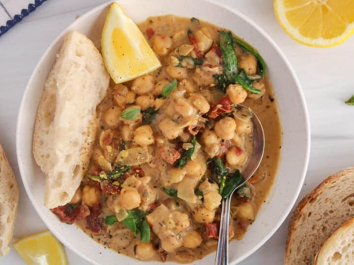 A spoon in a bowl of creamy vegan Tuscan chickpeas with a side of bread and lemon wedge.