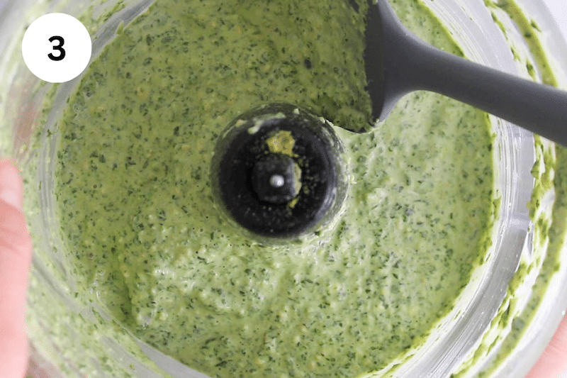 A spatula stirring in a food process with a creamy pesto sauce.