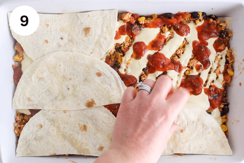 A hand placing tortillas over salsa in a large baking dish.