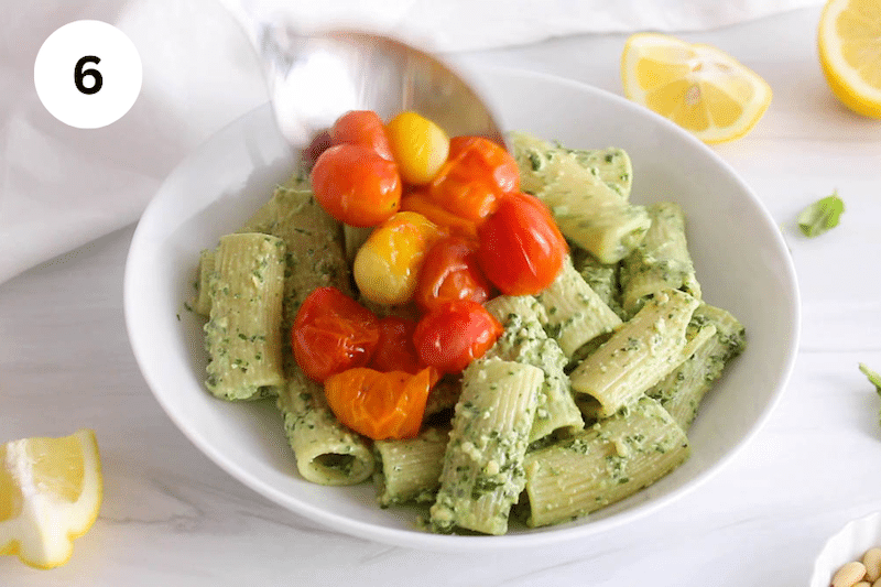 A spoon placing roasted cherry tomatoes on creamy pesto rigatoni.
