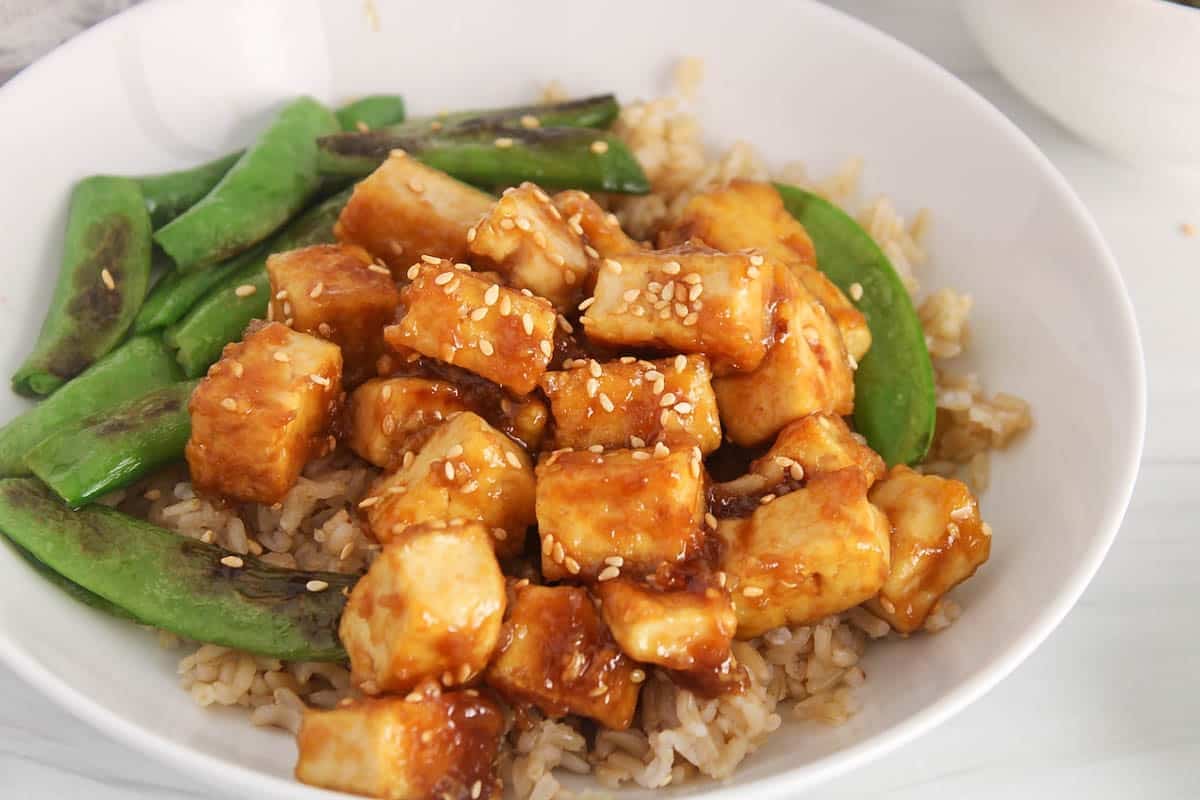 A bowl of sticky sesame ginger tofu over rice with a side of beans.