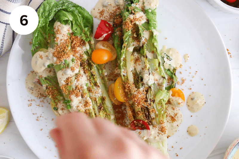 A plate with grilled romaine with dressing while a hand is sprinkling toasted breadcrumbs.