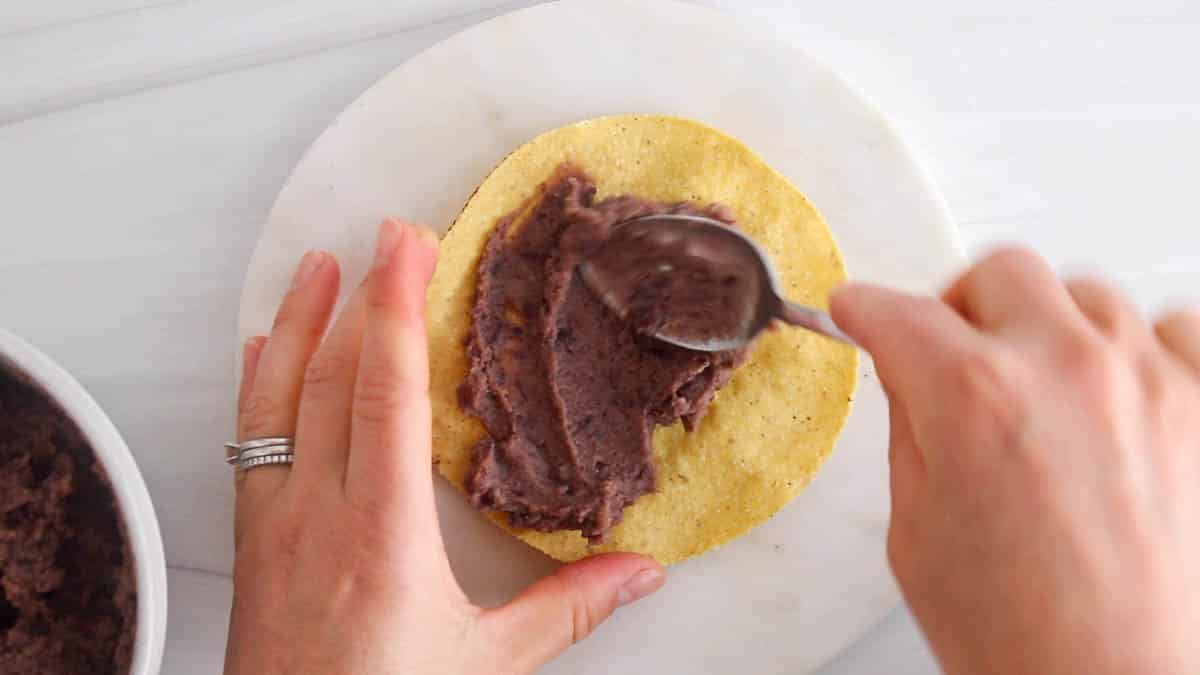 A spoon is spreading refried beans over a crispy tostadas.